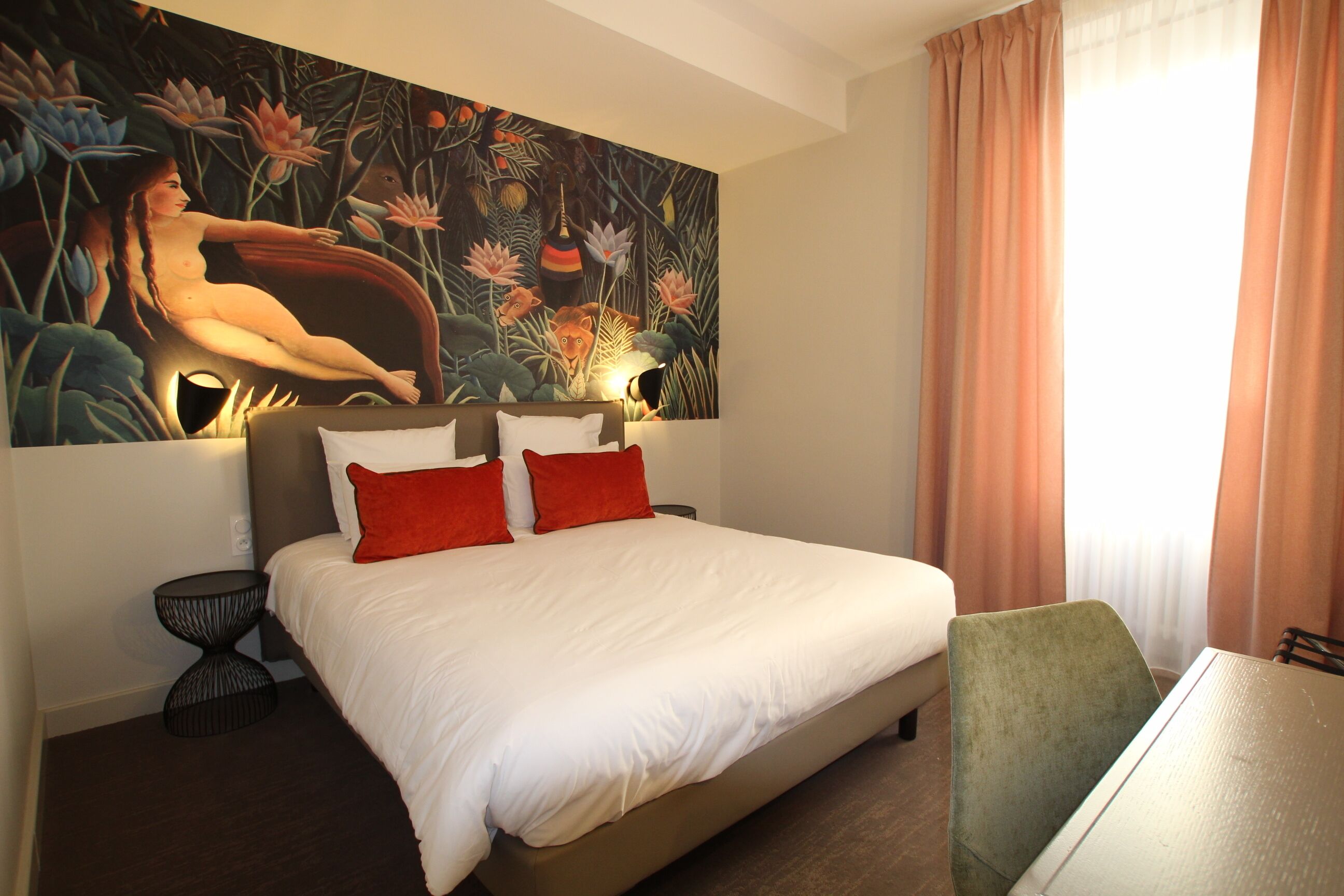 A hotel room with a large bed, two red pillows, a green chair, a small round table, and a wall painting.
