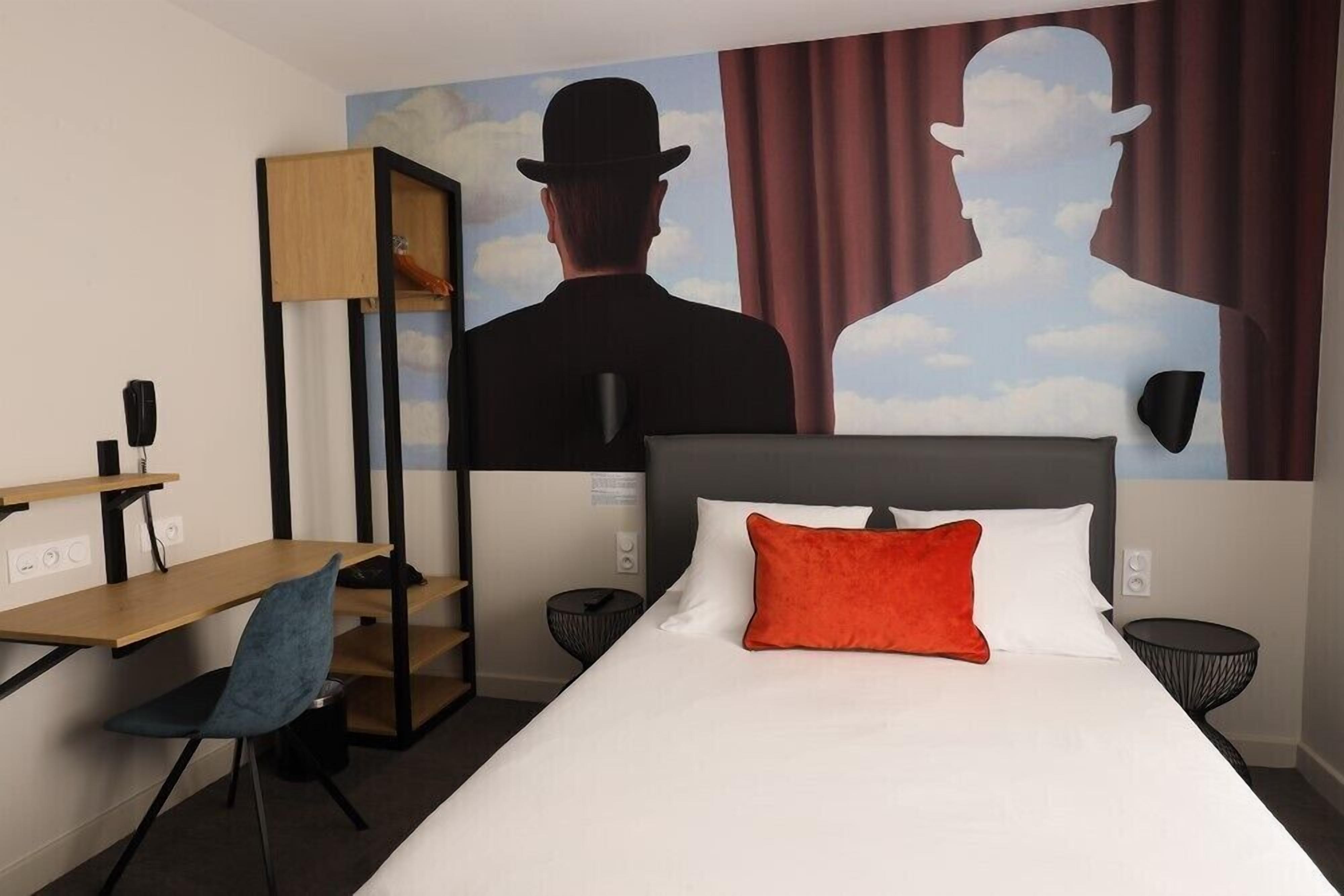 Une chambre d’hôtel comprenant un lit, un bureau, une chaise, une petite table et une fresque murale.