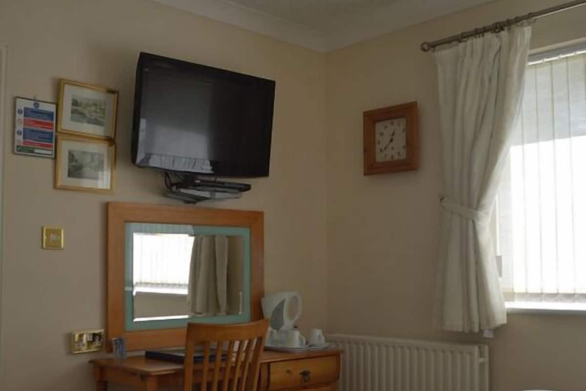 A hotel room with a bed, a desk with a chair, a television, a mirror, a clock, and a window with curtains.