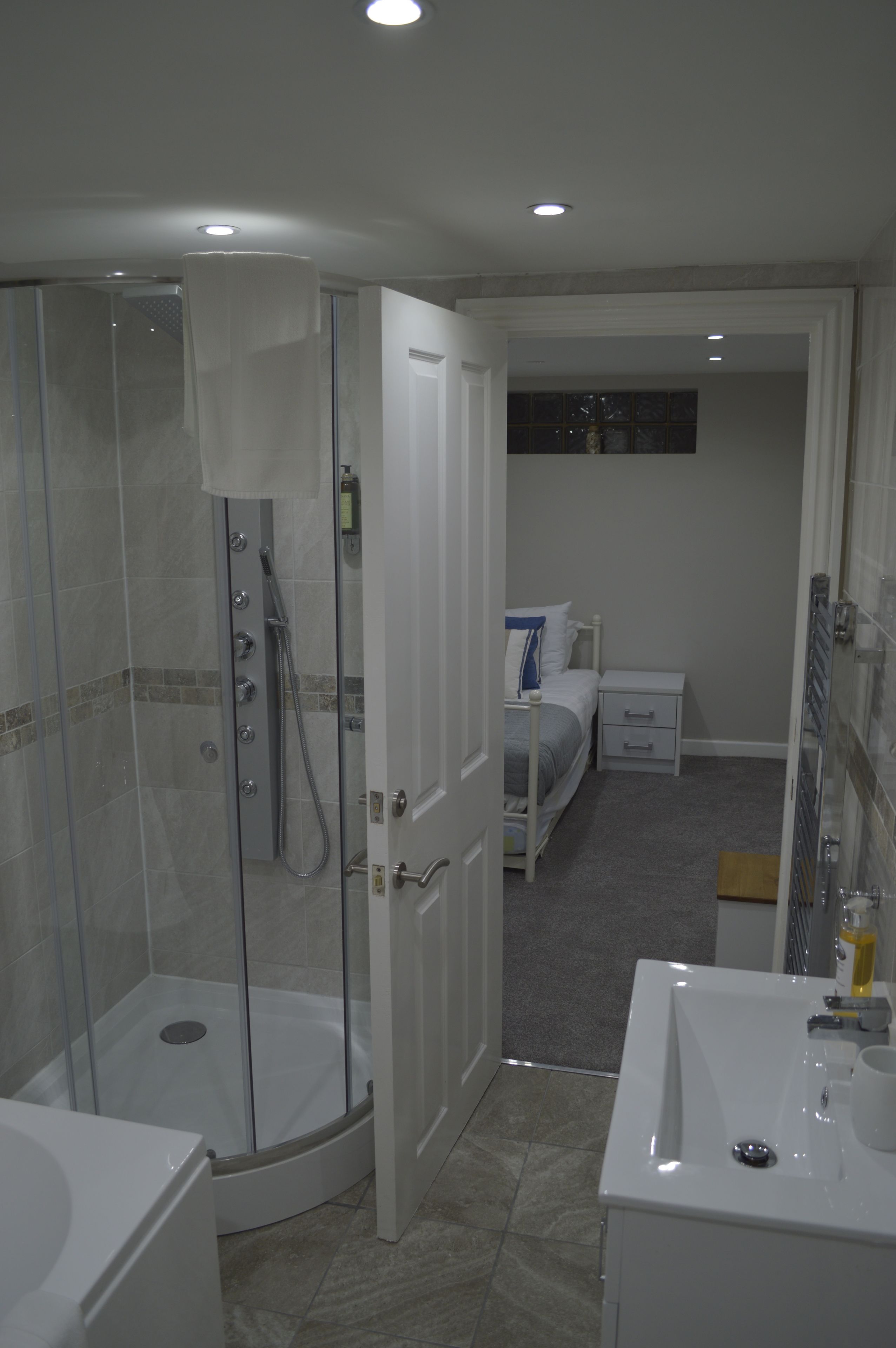 Un baño con ducha acristalada, un lavamanos blanco y una cama en la habitación contigua.