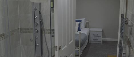 Un baño con ducha acristalada, un lavamanos blanco y una cama en la habitación contigua.