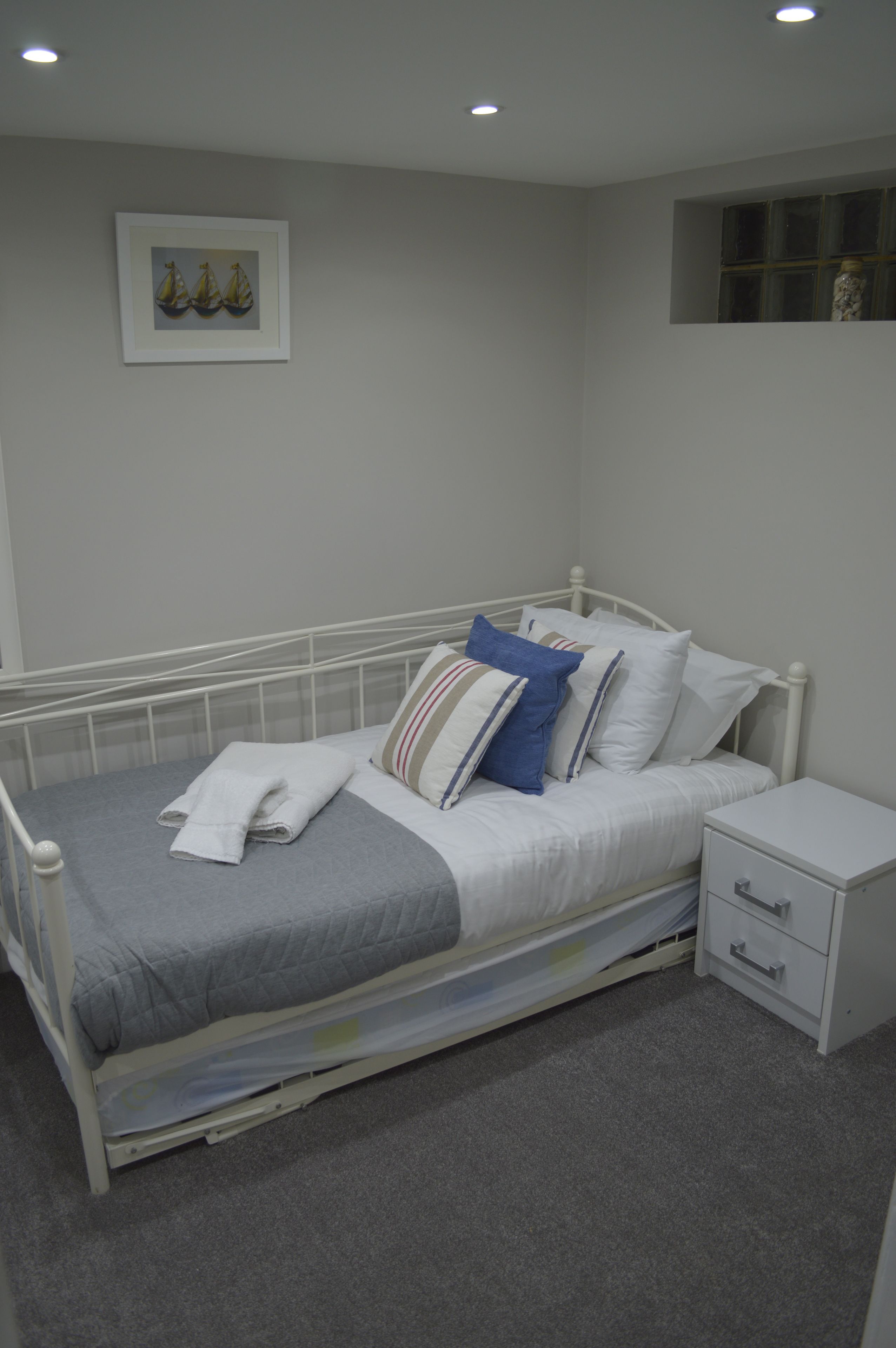 A neatly made bed with a grey blanket and pillows, a bedside table with drawers, and a framed picture on the wall.