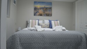 Een keurig opgemaakt bed met een grijze deken, kussens met strepen en een schilderij van een zonsondergang aan zee erboven.