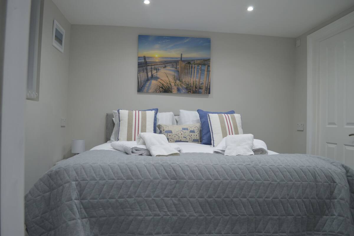 A neatly made bed with a gray quilt, pillows with striped patterns, and a beach sunset painting above it.