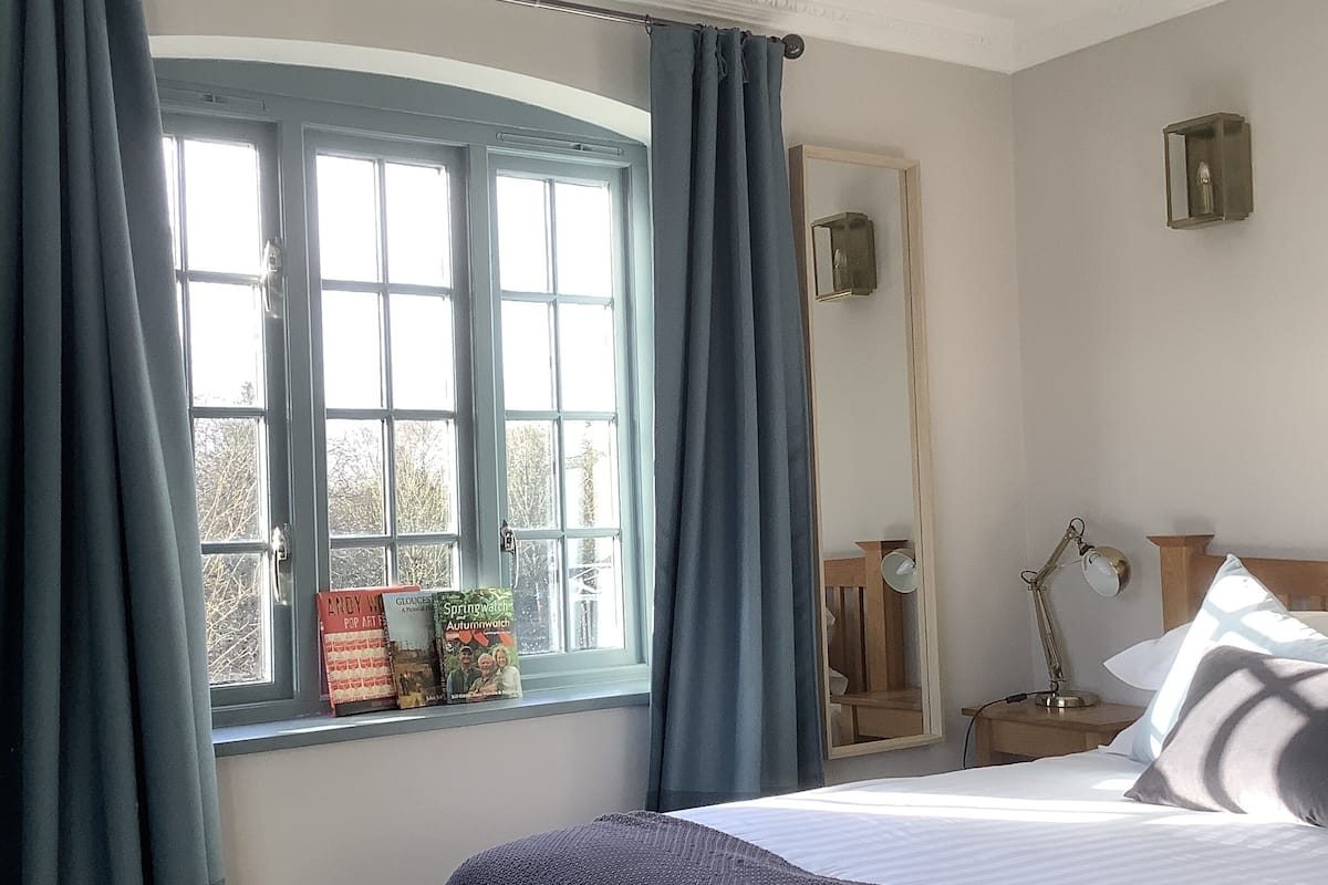 Une chambre avec une grande fenêtre, un lit recouvert d’une couverture bleue, une table de chevet avec une lampe et un miroir accroché au mur.