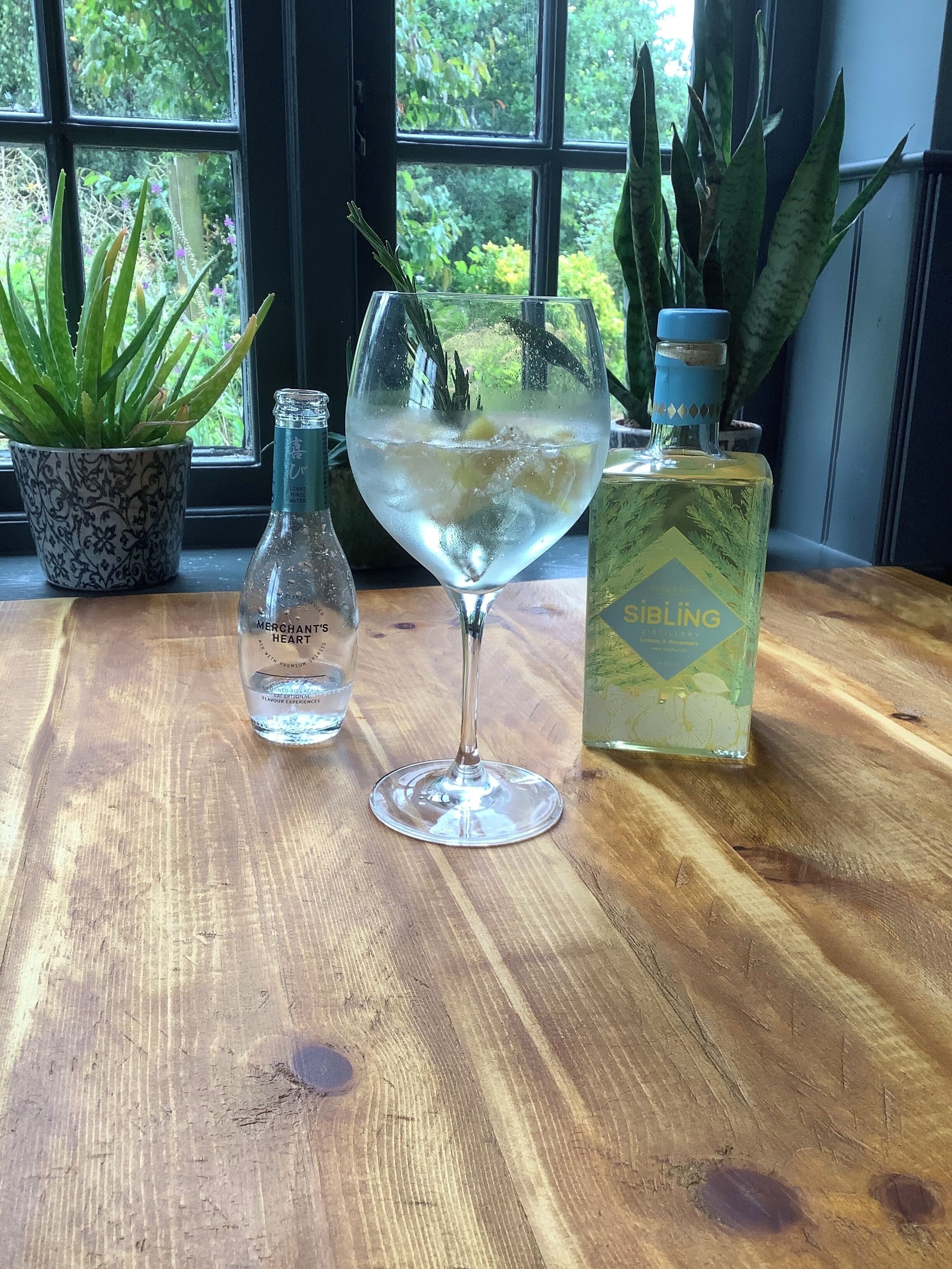 Una mesa de madera con un vaso de agua, una botella de Merchant's Heart y una botella de colonia Sibling.