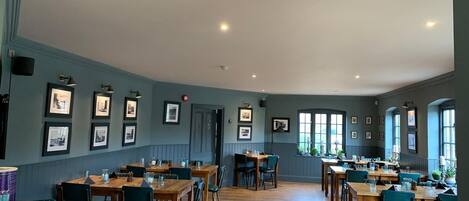A spacious dining area with multiple wooden tables and chairs, framed pictures on the walls, and large windows allowing natural light.