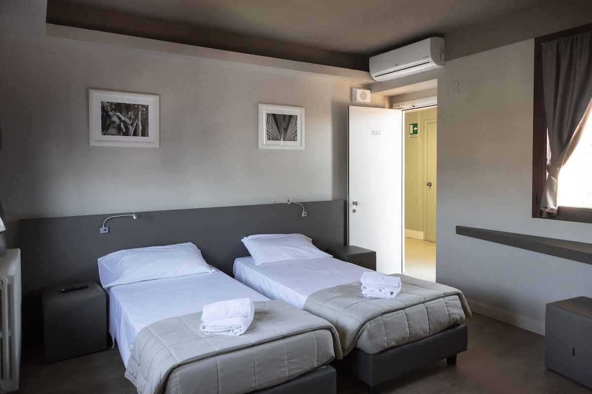 Una habitación de hotel con dos camas, una mesita de noche, un radiador y una ventana con cortinas.
