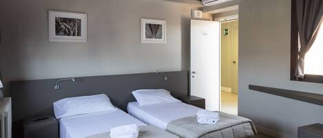 A hotel room with two beds, a bedside table, a radiator, and a window with curtains.