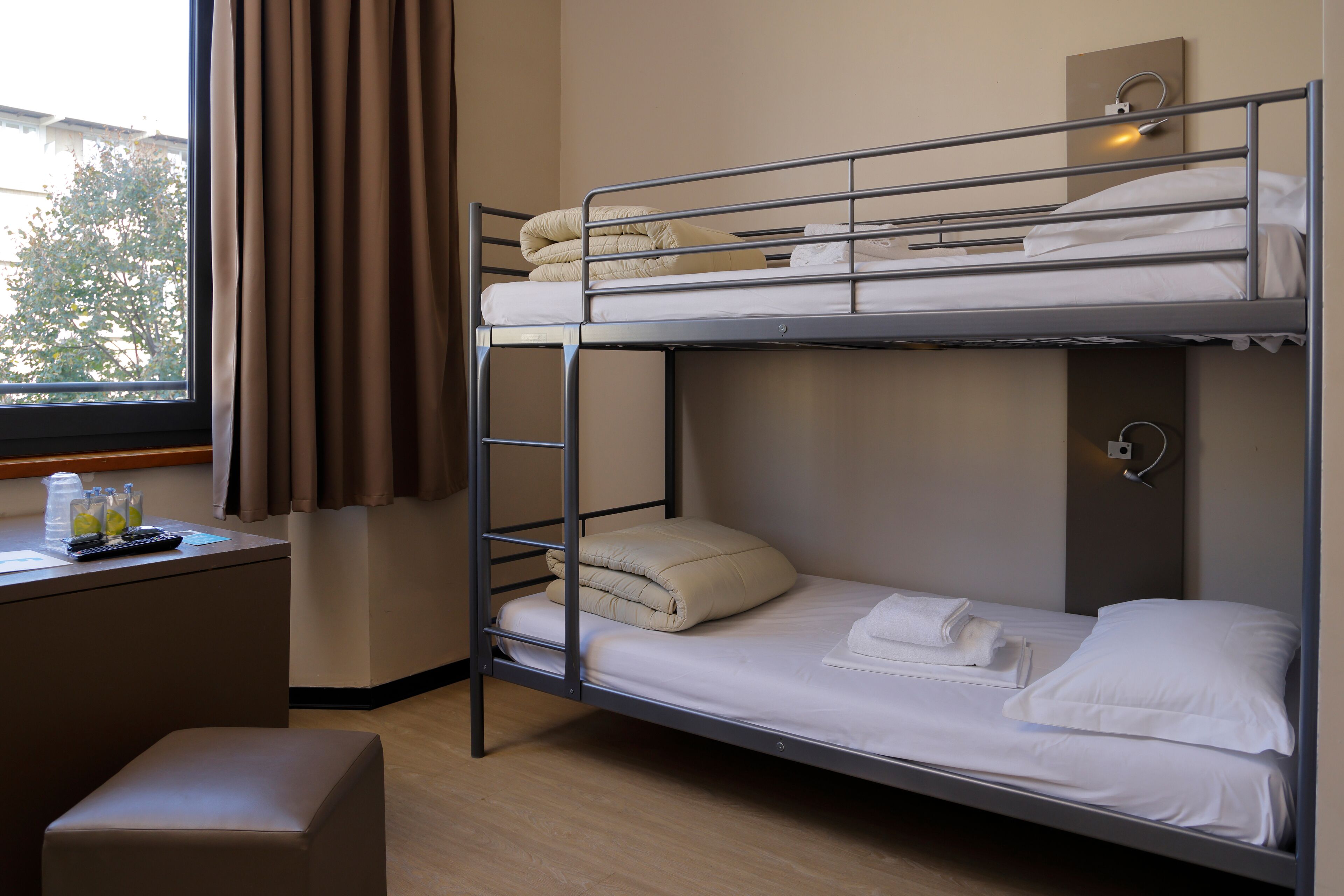 A bunk bed room with a desk, a window with curtains, and a bedside lamp.