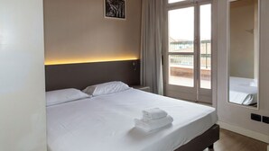 A hotel room with a large bed, a headboard with a light fixture, a window with curtains, and a balcony view.