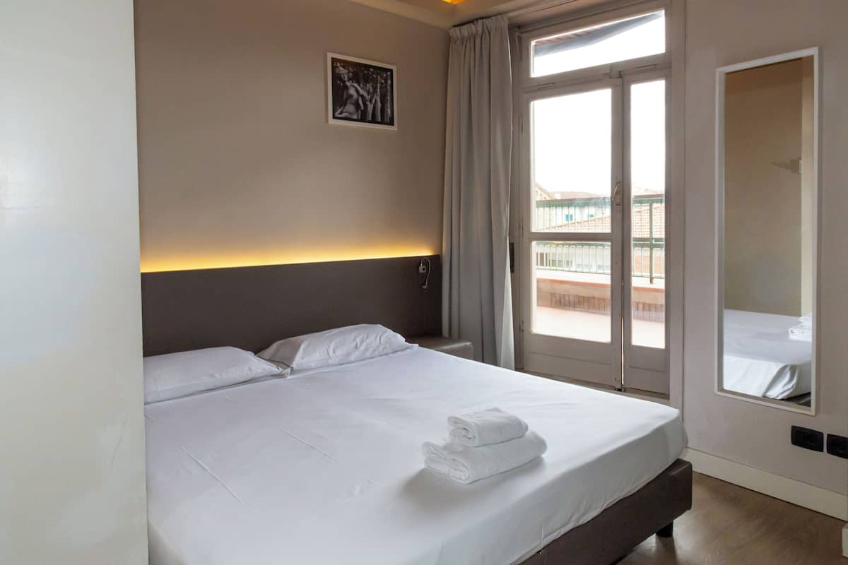 Habitación de hotel con una cama grande, cabecera con lámpara, ventana con cortinas y vistas al balcón.