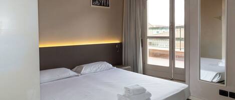 A hotel room with a large bed, a headboard with a light fixture, a window with curtains, and a balcony view.