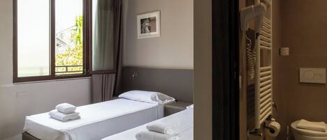 Habitación de hotel con dos camas individuales, una ventana con cortinas, un radiador, un inodoro y un cuadro en la pared.