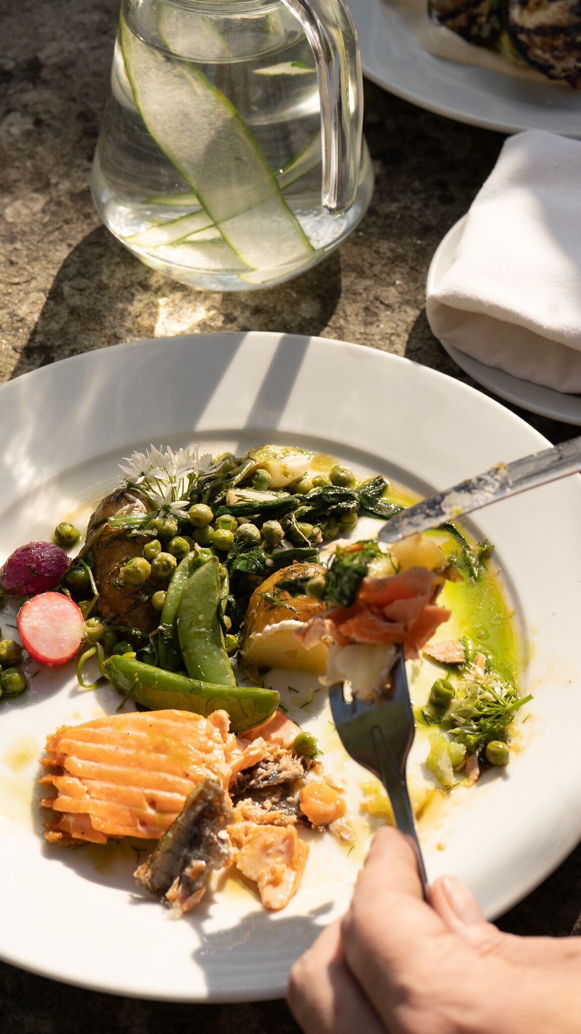 A plate of food with various vegetables and salmon, a glass pitcher with a lime slice, and a napkin on the table.