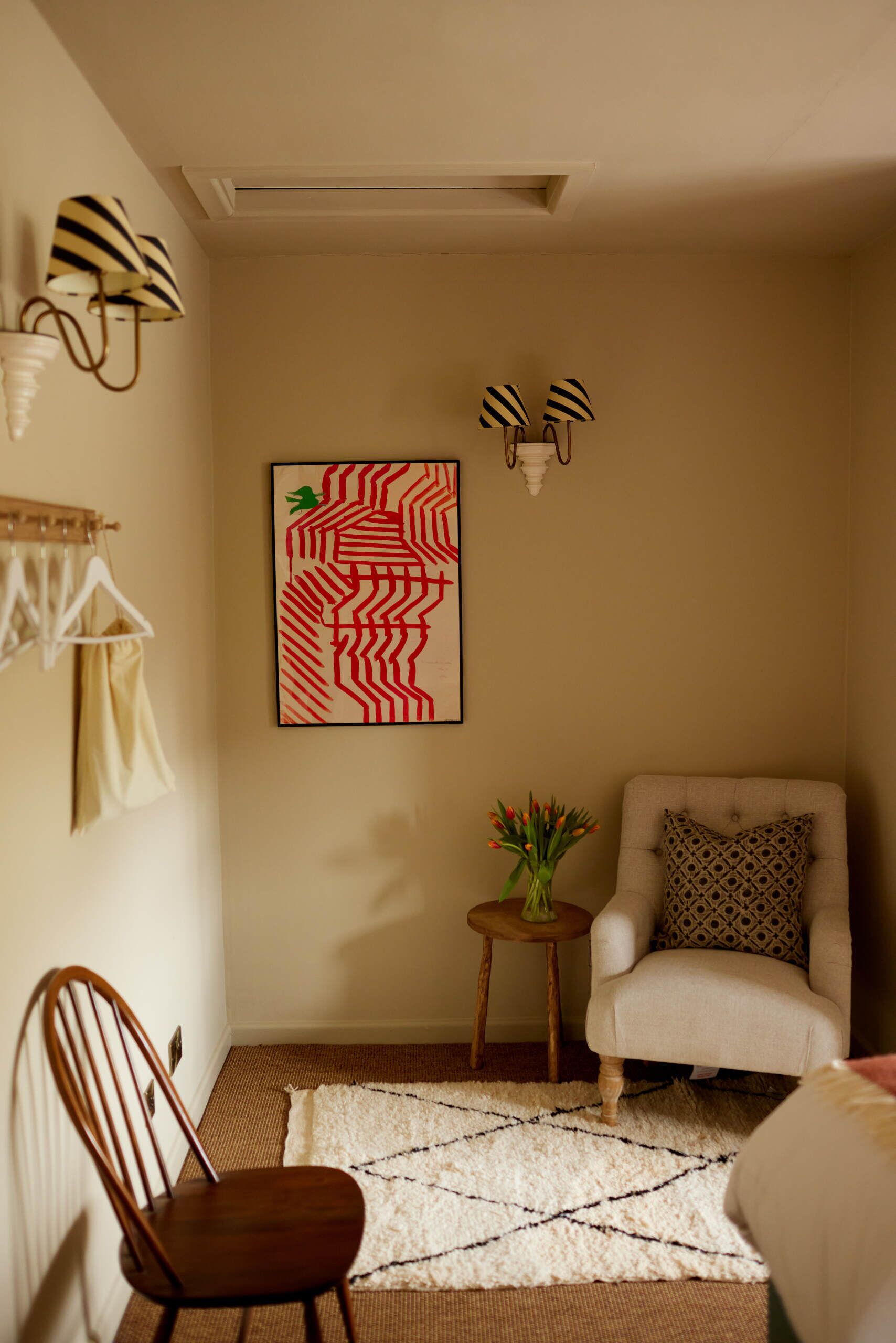 A room with a wooden chair, a white armchair, a patterned rug, a small side table with a vase, and a wall-mounted artwork.