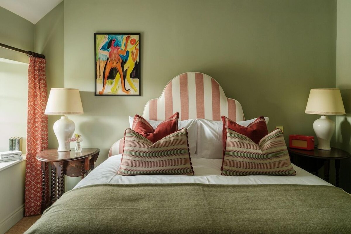 A bedroom with a striped headboard, two bedside lamps, a painting on the wall, and a window with curtains.