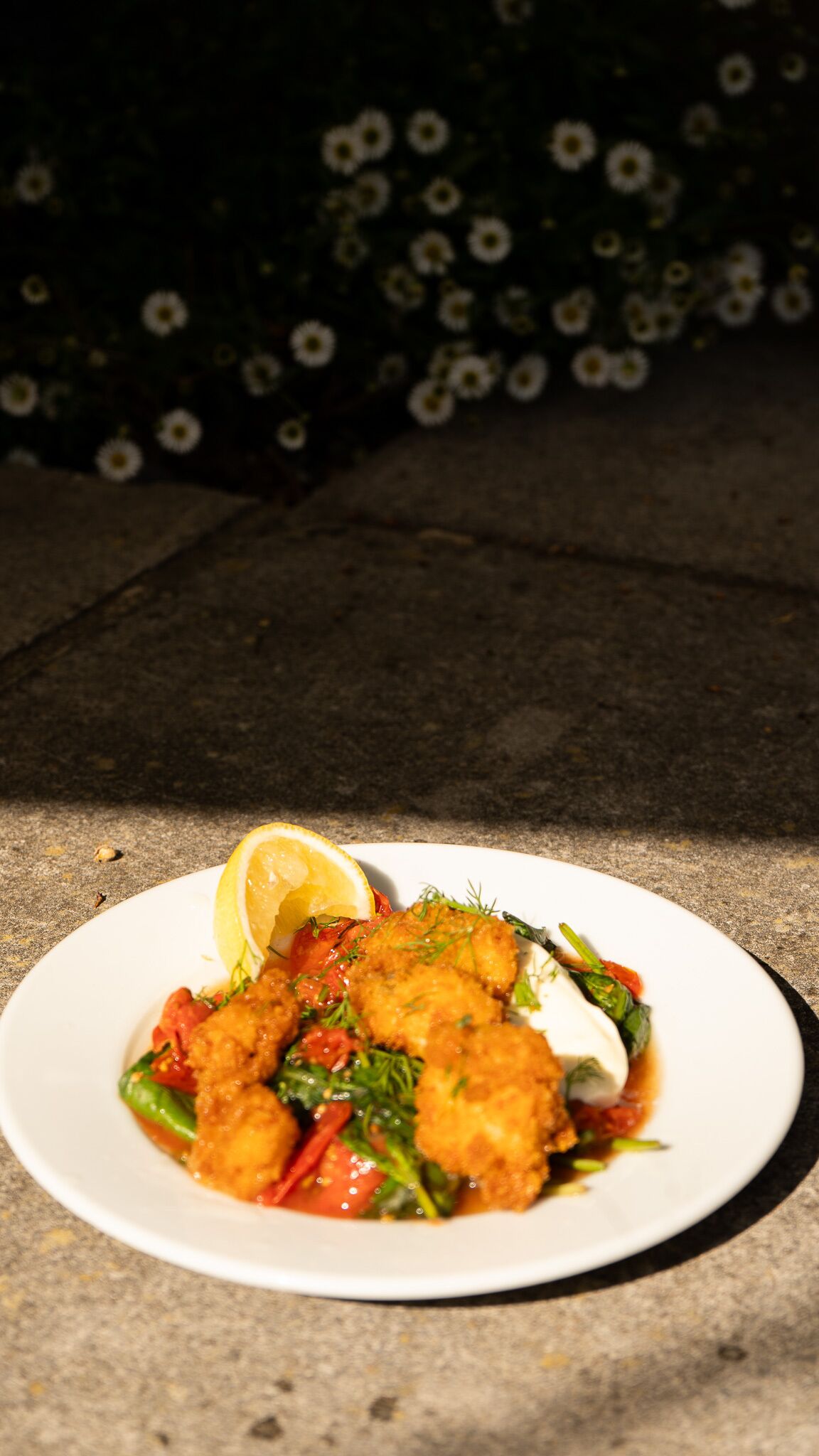 Un piatto di cibo con pesce impanato, verdure e spicchio di limone su un piatto bianco.