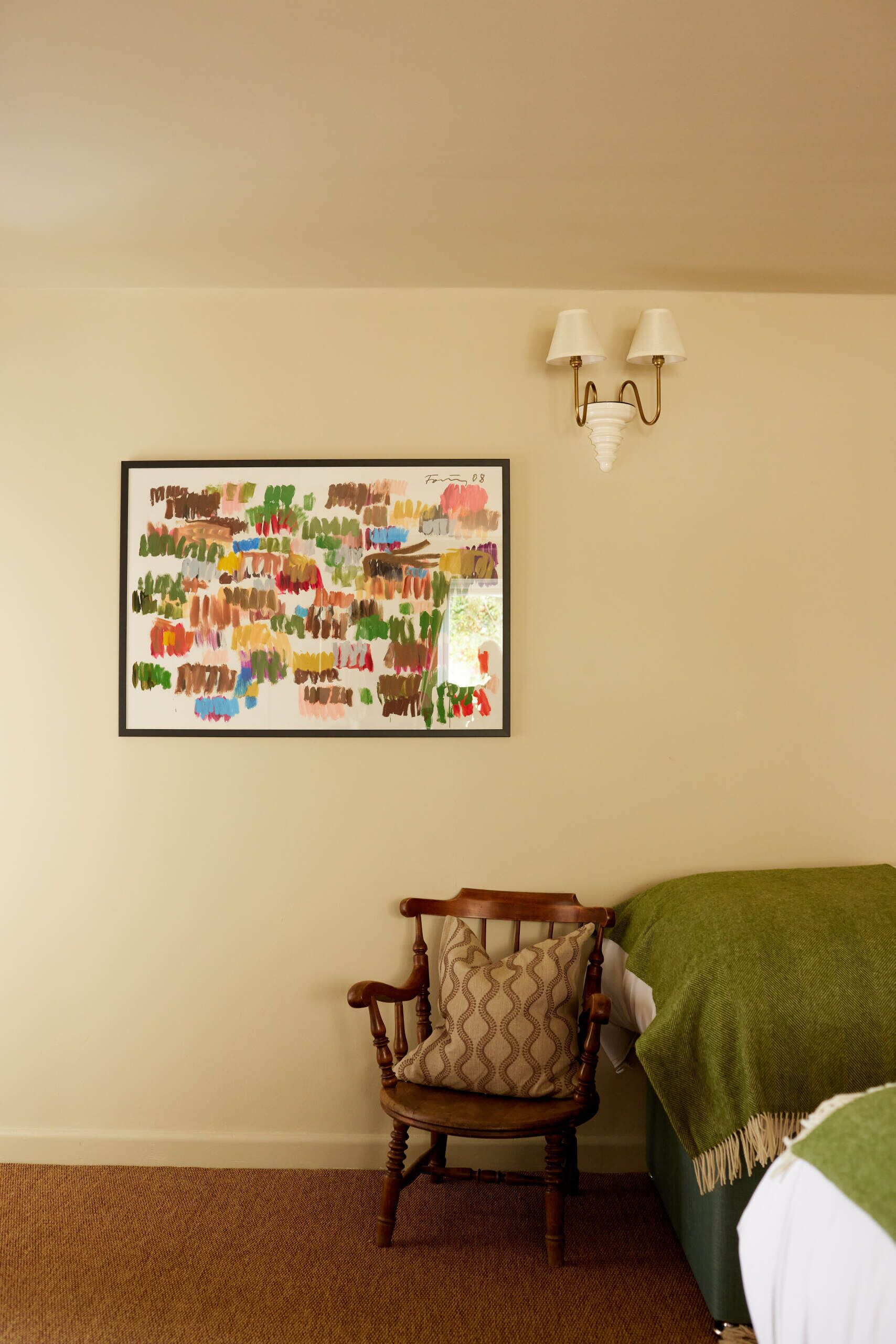 Una habitación con una silla de madera, una cama verde, una lámpara de pared y un cuadro abstracto colorido.