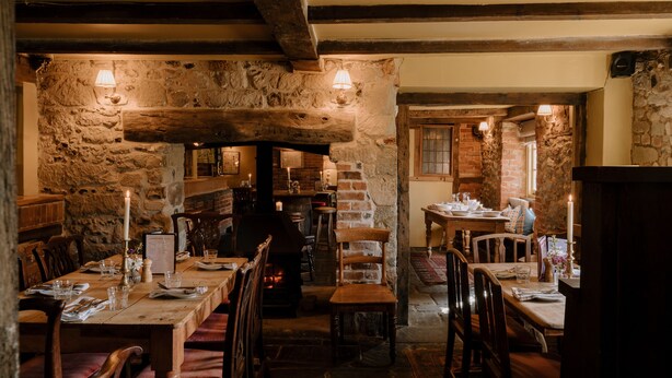 L’intérieur est de style rustique, avec des murs en pierre, des poutres en bois et une cheminée. Les tables sont dressées pour le repas, avec des bougies et des verres.