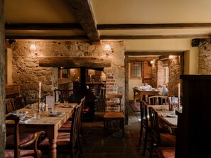 A rustic interior with stone walls, wooden beams, and a fireplace. Tables are set for dining with candles and glasses. - The Queen's Head (Salisbury)