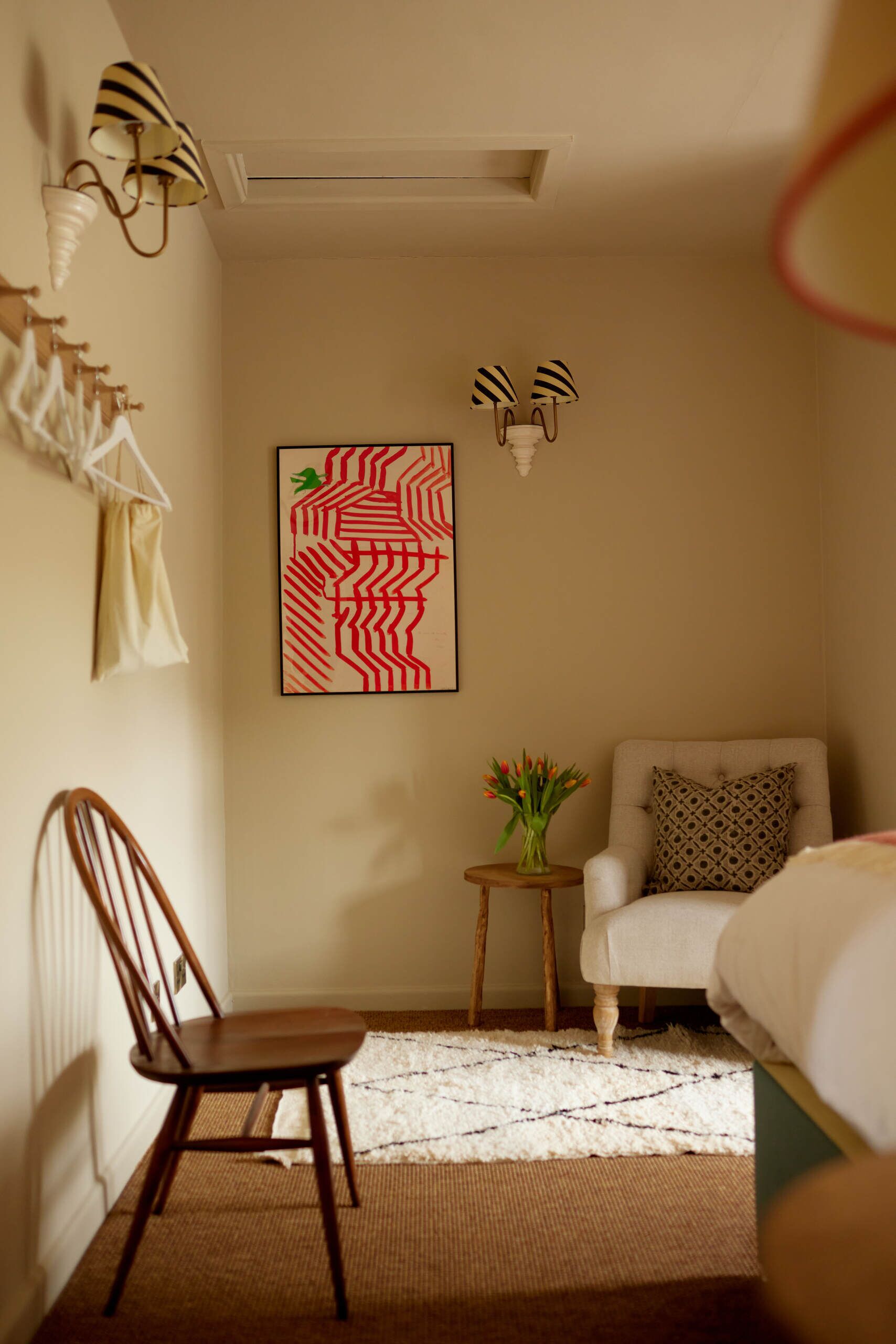 A small, cozy room with a chair, a small table with a vase of flowers, and a wall-mounted lamp.