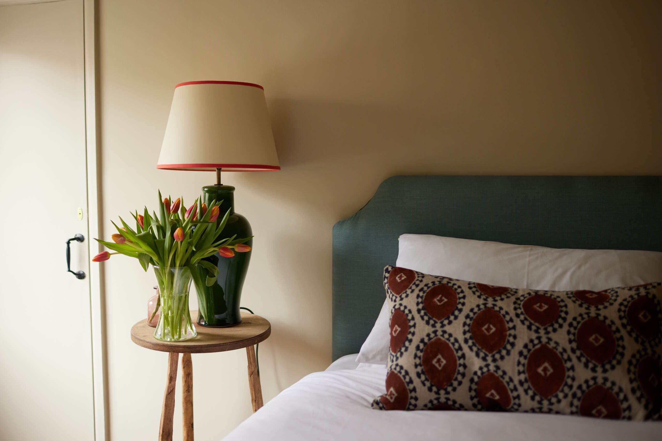 A neatly made bed with a patterned pillow and a bedside table with a lamp and a vase of flowers.