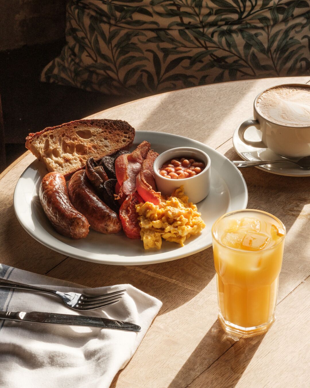 Un plato de desayuno con salchichas, tocino, tostadas, huevos revueltos, frijoles y un vaso de jugo de naranja.