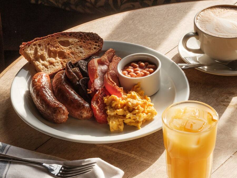 Un plato de desayuno con salchichas, tocino, tostadas, huevos revueltos, frijoles y un vaso de jugo de naranja.