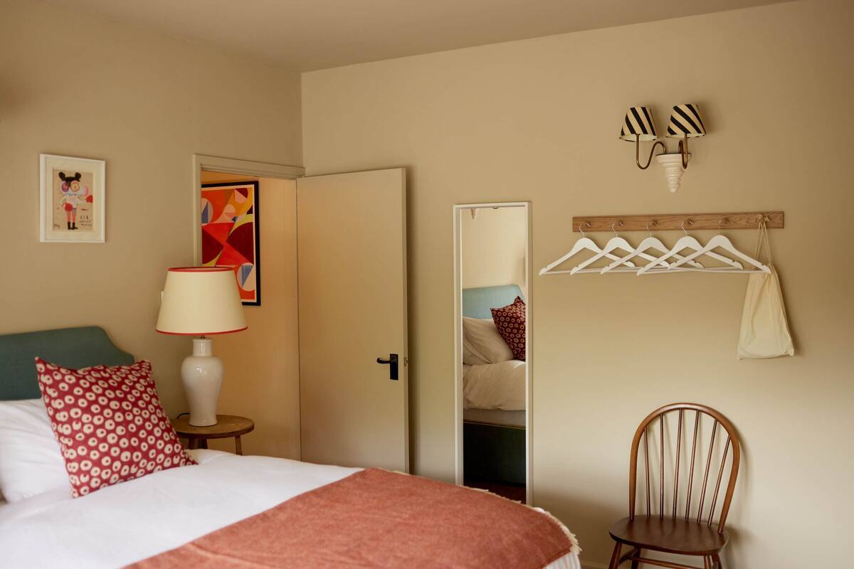 Una habitación de hotel con una cama, una silla, una lámpara, un espejo, un perchero y una pared decorada con obras de arte.