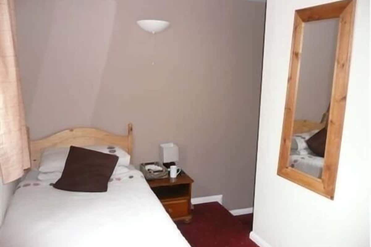 A small, neatly made bedroom with a single bed, a bedside table, a mirror, and a ceiling light.