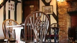 A traditional pub interior with exposed wooden beams, a brick fireplace, and a wooden chair with a metal backrest. - The Dog Inn (Halstead)