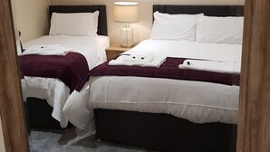 A hotel room with two beds, each with white linens and a dark headboard. There is a lamp on the nightstand between the beds. - The Dog Inn (Halstead)