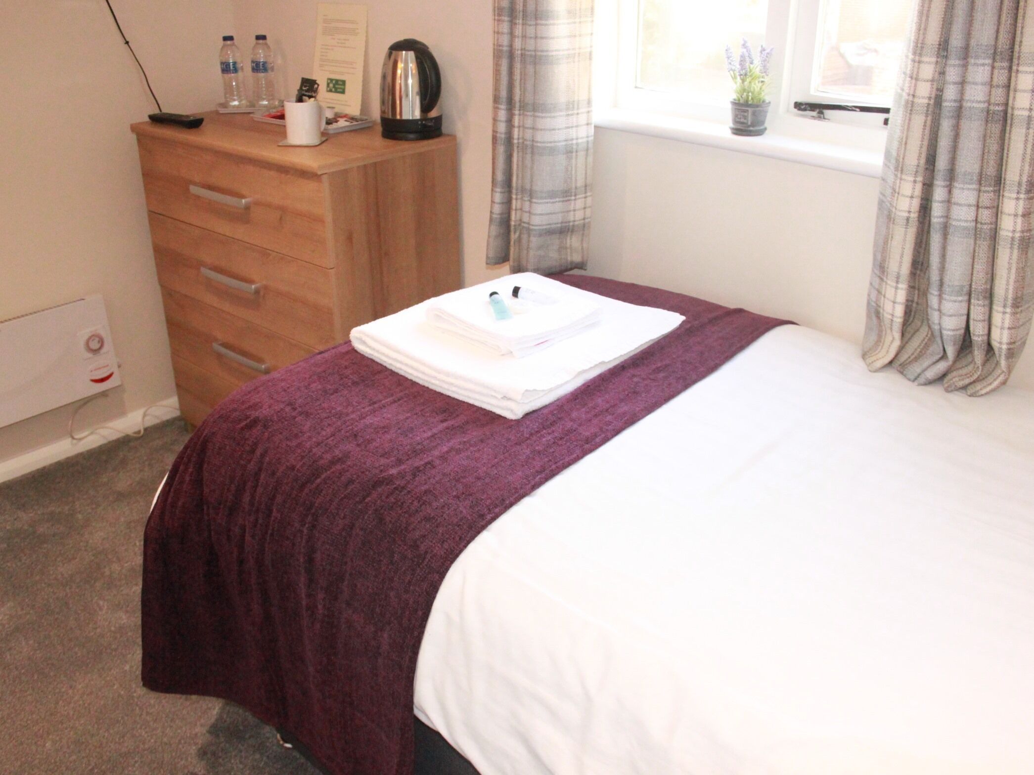 A hotel room with a bed, a wooden dresser, a window with curtains, and a radiator.