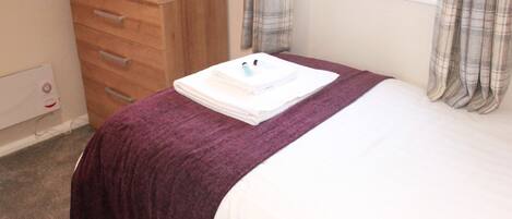 A hotel room with a bed, a wooden dresser, a window with curtains, and a radiator.