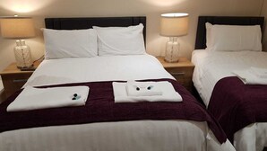 A hotel room with two beds, each with white linens and pillows. There are two bedside tables with lamps and a decorative vase on one. The room has a simple, clean design with a dark headboard. - The Dog Inn (Halstead)