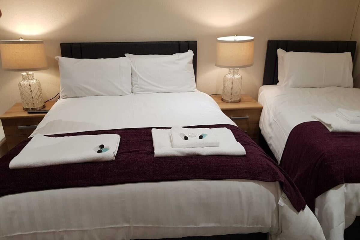 A hotel room with two beds, each with white linens and pillows. There are two bedside tables with lamps and a decorative vase on one. The room has a simple, clean design with a dark headboard.