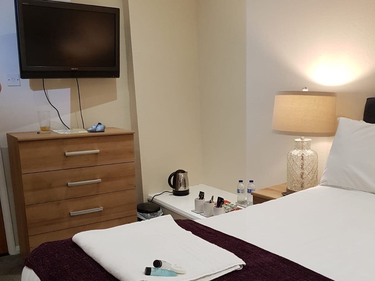 A hotel room with a bed, a wooden dresser, a television mounted on the wall, a nightstand with a lamp, and a kettle.