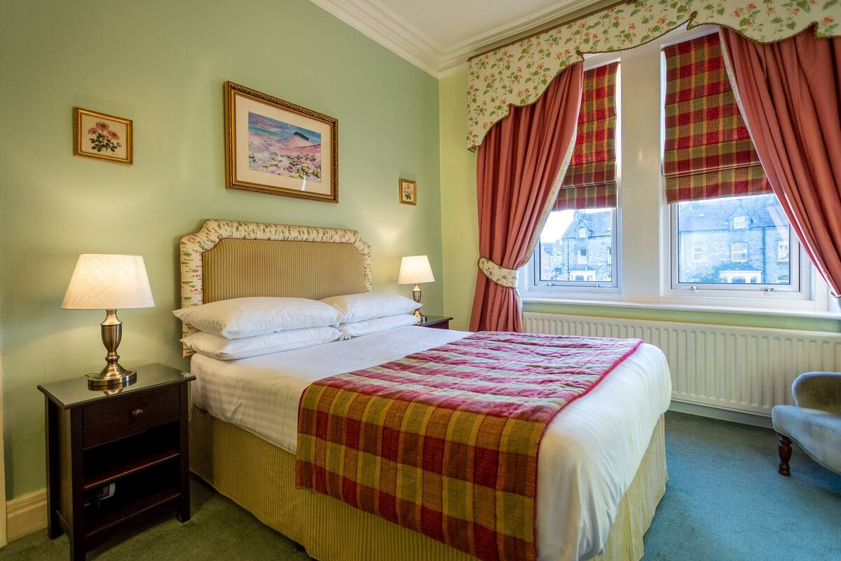 Un dormitorio ordenado con una cama, dos lámparas de noche, una mesita de noche, una silla y una ventana con cortinas.