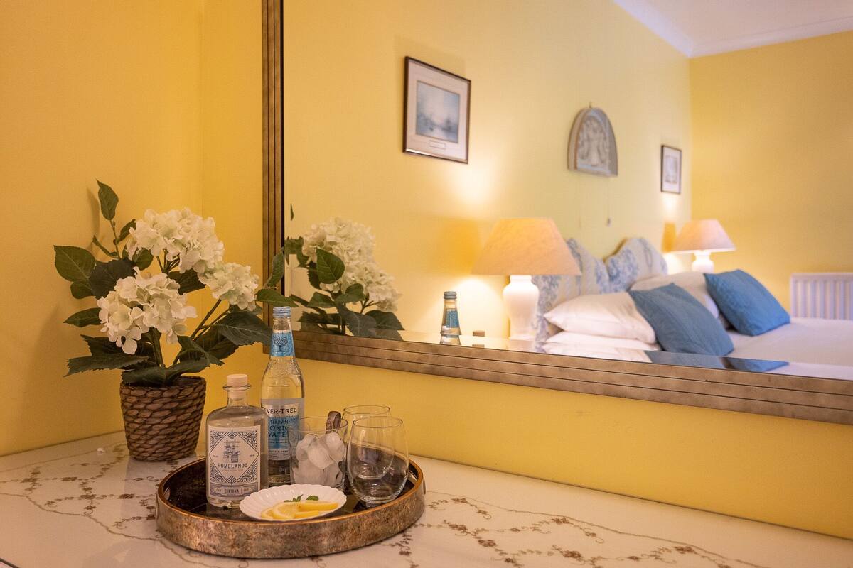 Una habitación de hotel con una cama, una mesita de noche con un bandeja de artículos de aseo, un espejo que refleja la habitación y una pared amarilla.