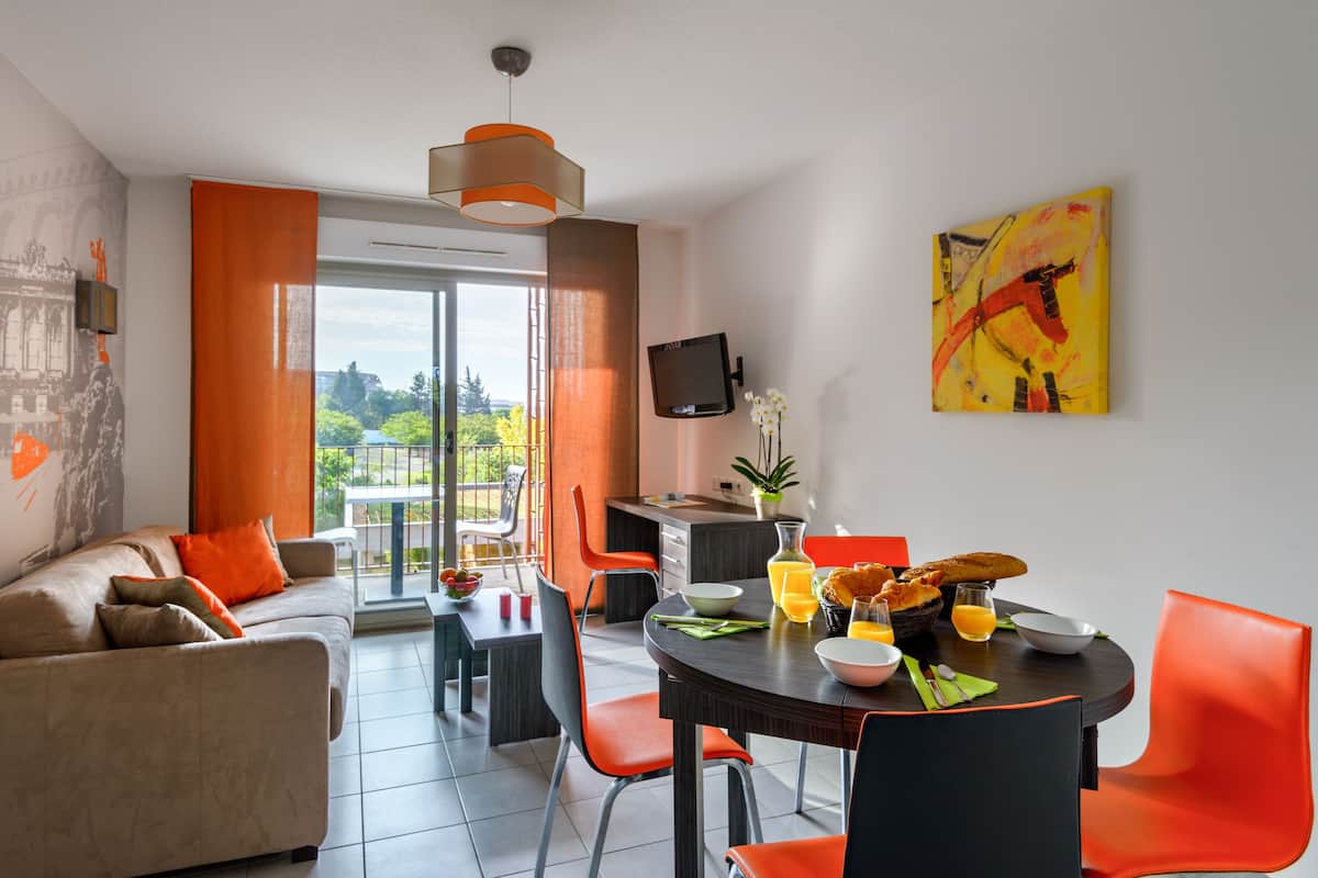 Una sala de estar moderna con sofá, mesa de comedor y sillas. Hay un balcón con vistas a zonas verdes.