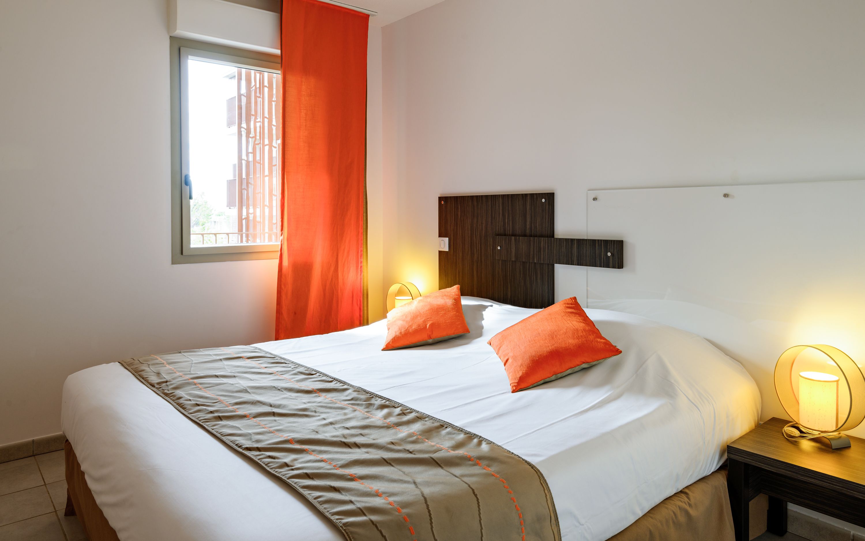 Une chambre d’hôtel comprenant un lit, deux oreillers, une table de chevet avec une lampe, une fenêtre avec des rideaux et une tête de lit fixée au mur.
