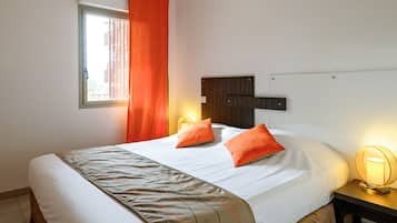 Une chambre d’hôtel comprenant un lit, deux oreillers, une table de chevet avec une lampe, une fenêtre avec des rideaux et une tête de lit fixée au mur.