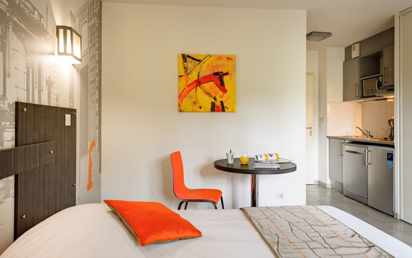 Una habitación de hotel moderna con una cama, una pequeña mesa con una silla, una zona de cocina y un cuadro en la pared.