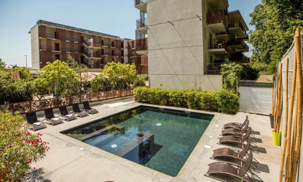 Una piscina amb gandules, envoltada de vegetació i edificis moderns.