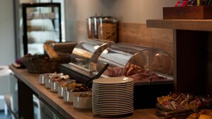 Mostrador de buffet con diversas opciones de comida, como carnes, quesos y pasteles, junto con utensilios y platos apilados.