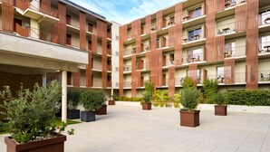 Un edificio residencial de varios pisos con balcones, un patio con plantas en macetas y un cielo despejado.