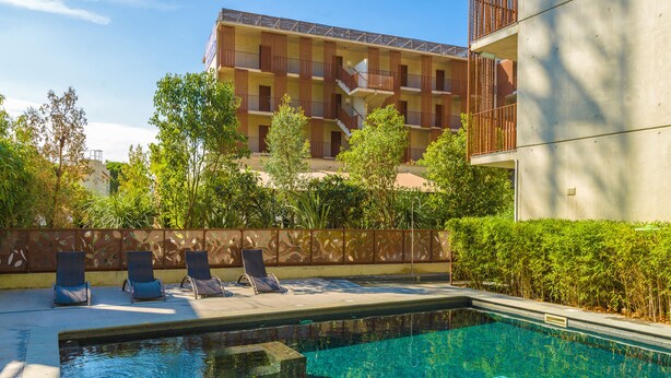 Una piscina con sillas de descanso, un edificio con balcones y abundante vegetación.