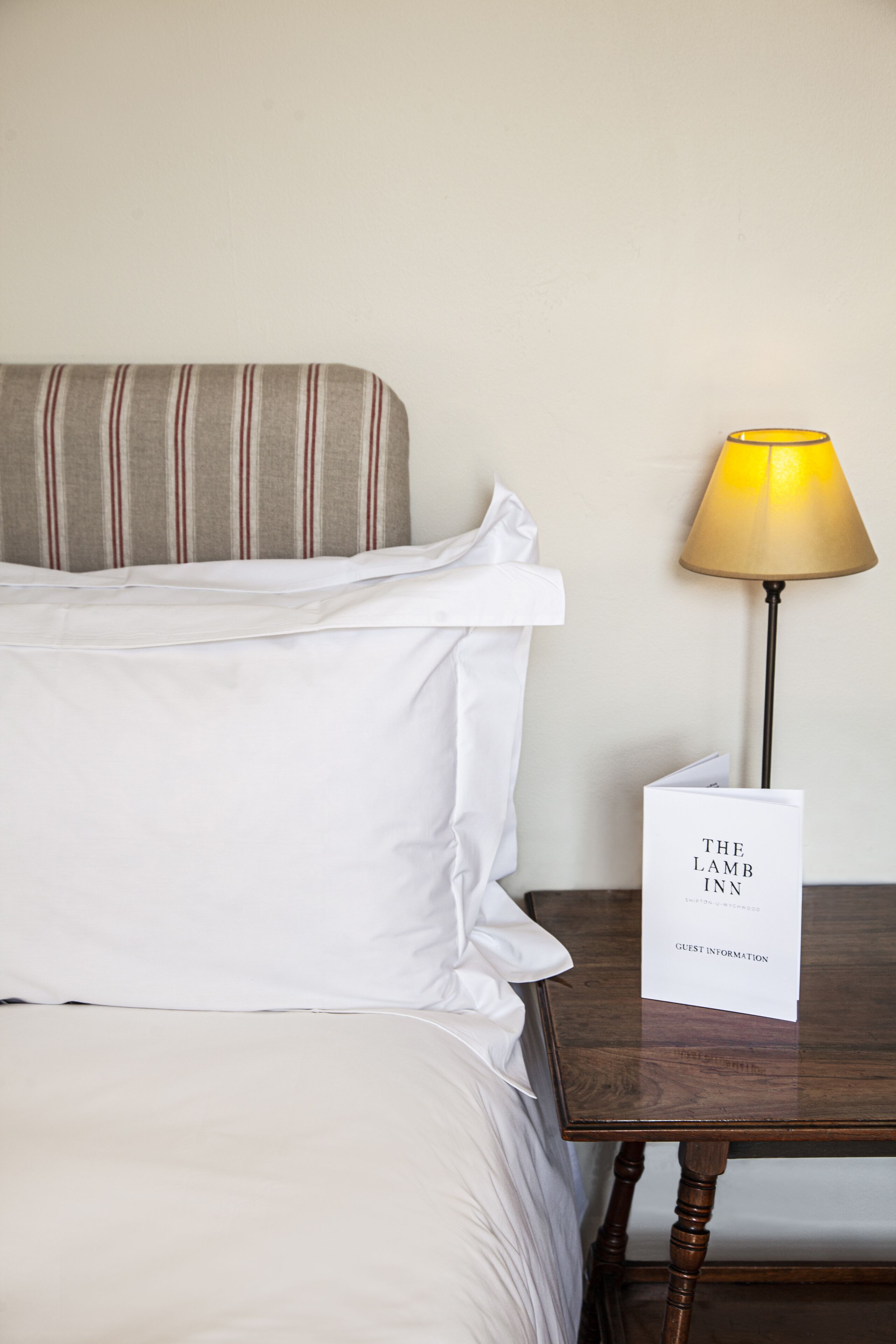 A neatly made bed with white linens, a striped pillow, and a table with a lamp and a guest information card.