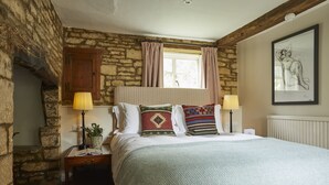 A bedroom with a stone wall, a bed with patterned pillows, a nightstand with a lamp, and a framed artwork on the wall. - The Lamb Inn (Chipping Norton)